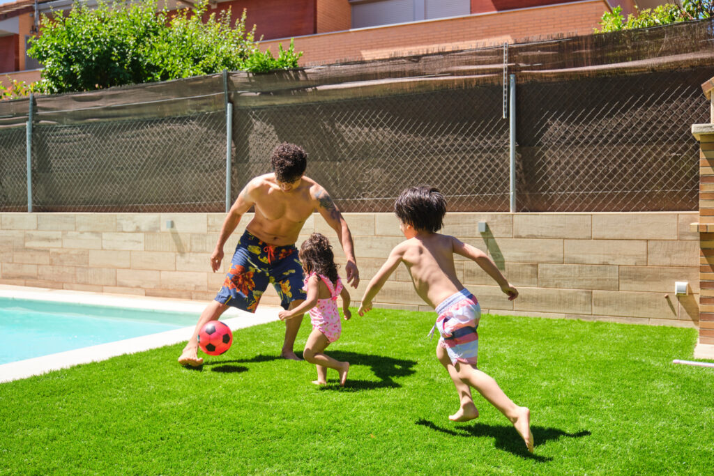 Un hombre y sus dos hijos jugando al fútbol en un jardín con piscina